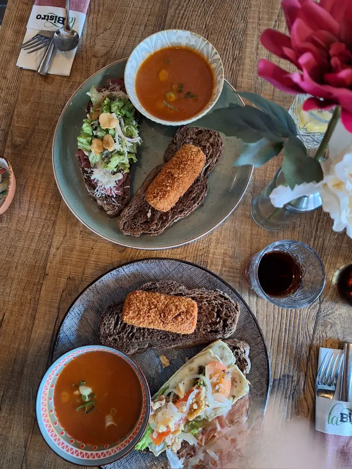 Van bovenaf: twee borden met soep, een kroket op bruinbrood en belegde boterhammen op een houten tafel